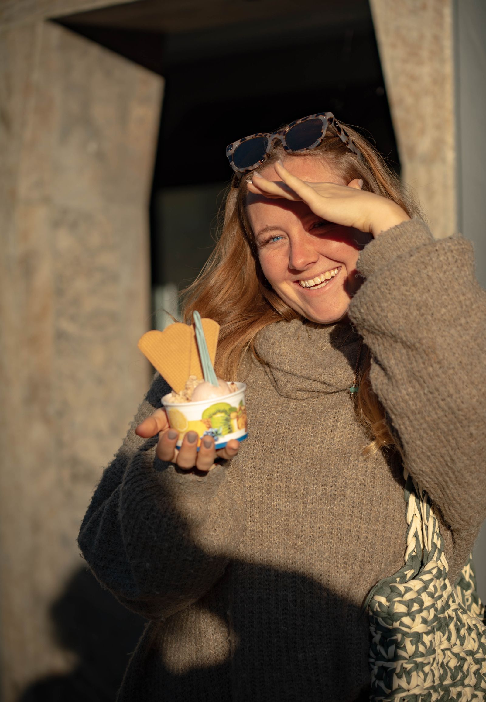 Lachende Frau mit Eisbecher schirmt die Augen gegen die tiefe Sonne ab