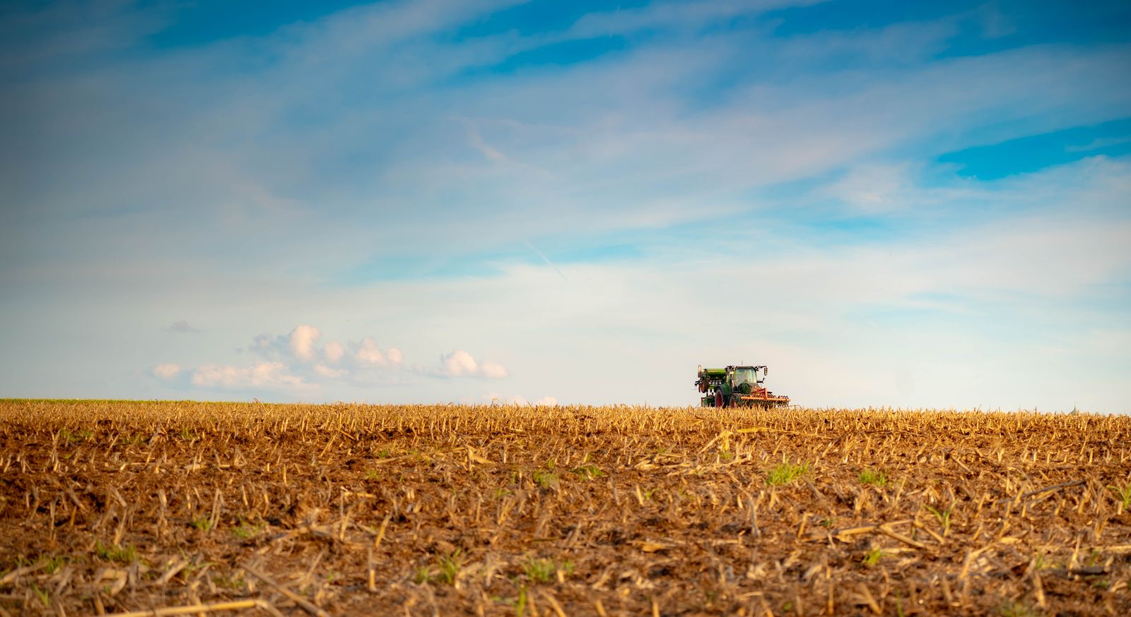 Weites Stoppelfeld unter blauem Himmel mit einem kleinen Traktor am Horizont