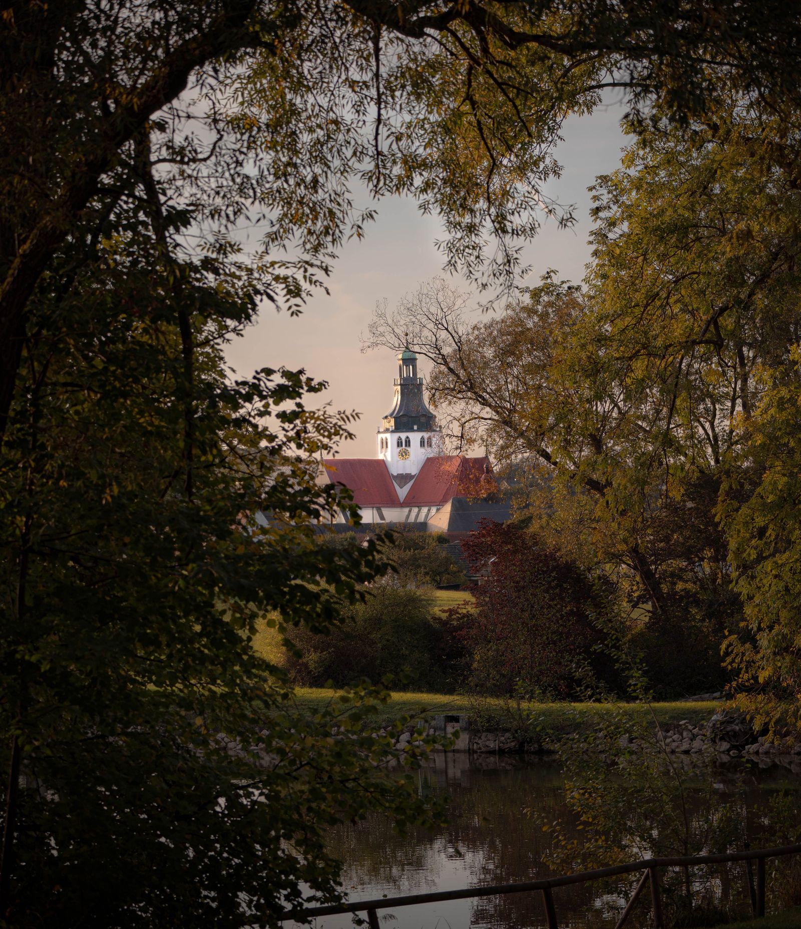 Kirchturm durch einen Rahmen aus Bäumen, davor ein Weiher